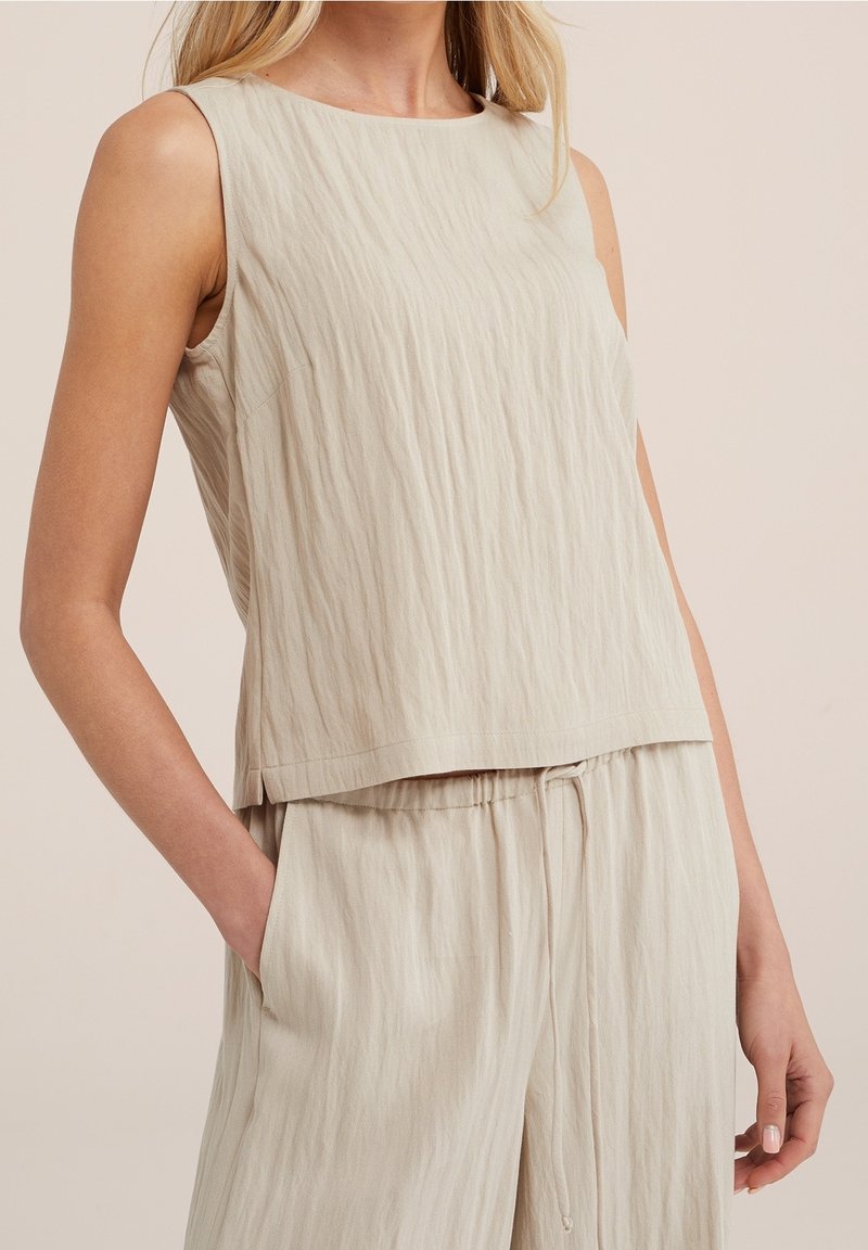 Haut sans manches beige clair avec un design texturé et ondulé. Il présente un col rond et un ourlet droit, associé à un pantalon ample.