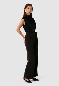 Svart satin utenermet topp med høy hals, kombinert med høylivete svarte bukser og svarte slingbacks med spiss tå.