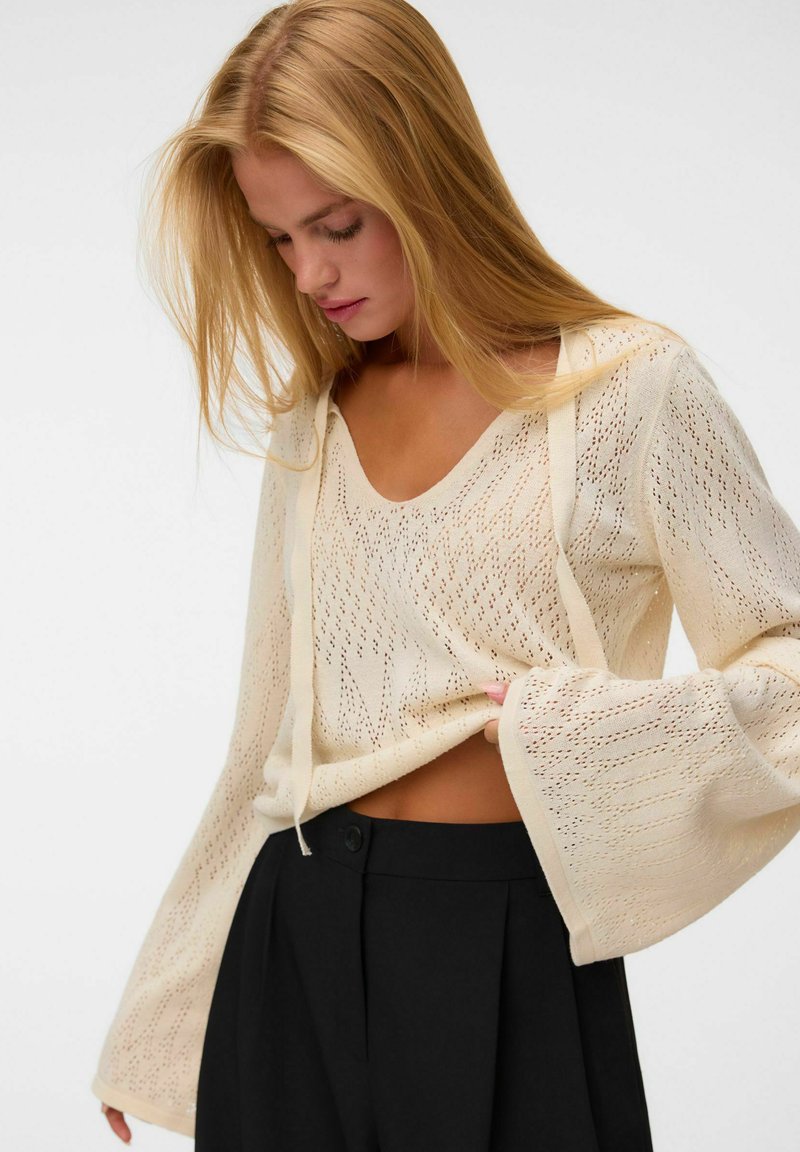 Femme blonde regardant vers le bas, portant un haut en maille crème à manches longues évasées et un pantalon noir taille haute.
