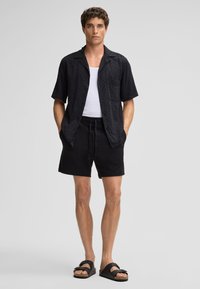Zwarte korte mouwen overhemd met textuurpatroon, gecombineerd met zwarte shorts en zwarte sandalen, staande tegen een neutrale achtergrond.