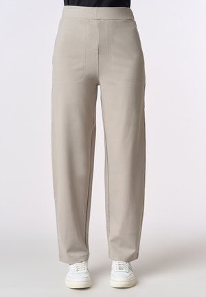 Pantaloni a gamba larga color beige chiaro con una texture liscia, dotati di tasche anteriori e dettagli cuciti, abbinati a sneakers bianche.