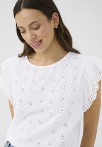 Top en coton blanc avec broderie florale en œillets, décolleté rond et manches courtes à volants. Présente une surface texturée avec de délicats découpes.