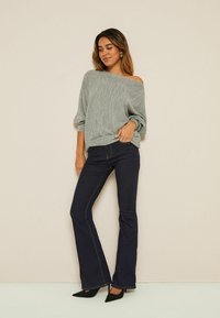 Pullover a maglia grigia chiara, testurizzata e senza spalle abbinato a jeans a zampa d'elefante blu scuro e tacchi neri a punta, esposto su uno sfondo neutro.