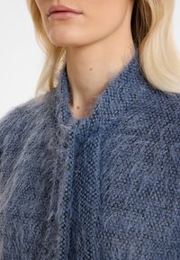 Flauschige blaue Strickjacke mit weicher Textur, hochgeschlossen und leicht lockerer Passform. Kabelstrickdetails sind sichtbar.