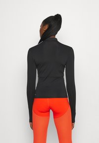 Kobieta z długim warkoczem w kucyku, ubrana w dopasowaną czarną bluzę z długim rękawem i jasnopomarańczowe teksturowane legginsy, stojąca plecami.