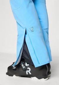 Hemelsblauwe sneeuwbroeken met ritssluitingen, voorzien van een logo-patch, gedragen met zwarte skischoenen met witte letters op de zijkanten.
