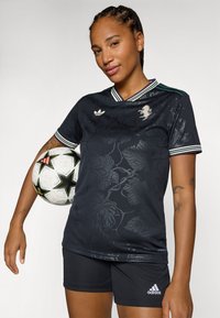 Maglia da calcio nera con motivo botanico, colletto a righe bianche e verdi, abbinata a pantaloni corti neri e holding un pallone da calcio decorato.