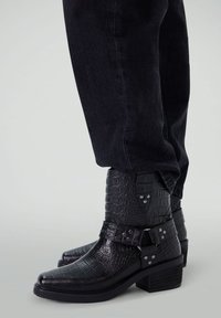 Bottines noires en simili cuir avec une texture crocodile, un bout pointu, des accents de boucle et un talon empilé. Associées à un jean noir.