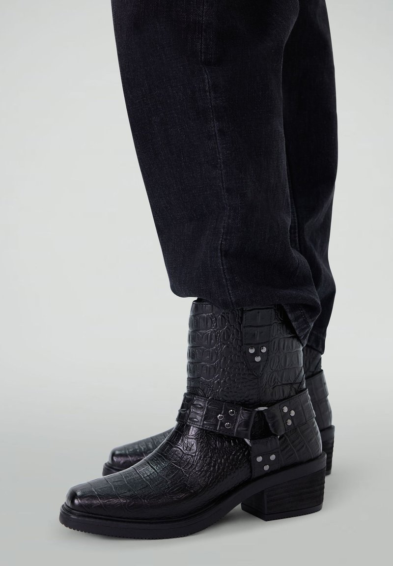 Bottines noires en simili cuir avec une texture crocodile, un bout pointu, des accents de boucle et un talon empilé. Associées à un jean noir.