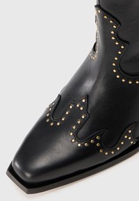 Stivale da cowboy in pelle nera con punta affilata, decorato con borchie dorate e ritagli ondulati lungo la parte superiore per un design intricato.