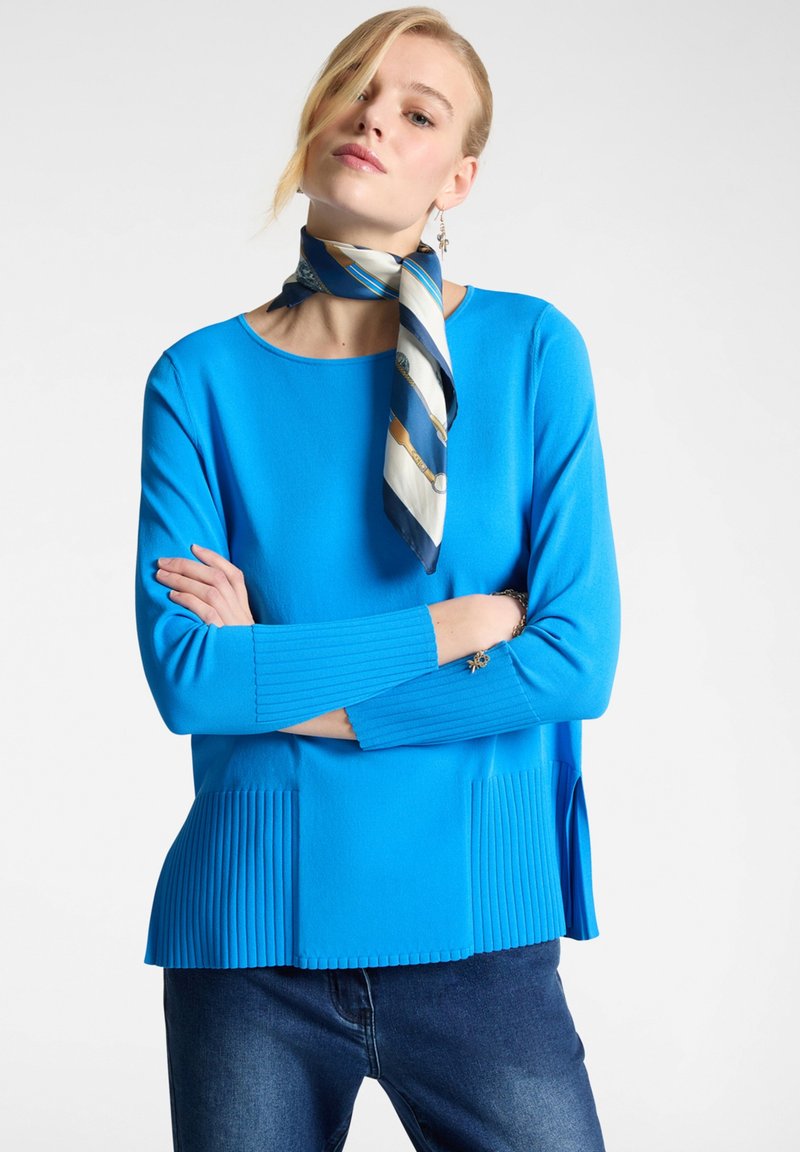 Blue ribbed long-sleeve top with pleated hem and side slits. Paired with a patterned scarf and denim jeans. Smooth texture.