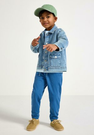 Nuori poika vihreä lippalakki päässään, farkkutakki, siniset housut ja vaaleanruskeat kengät, osoittaa molemmilla käsillään eteenpäin yksiväristä taustaa vasten.