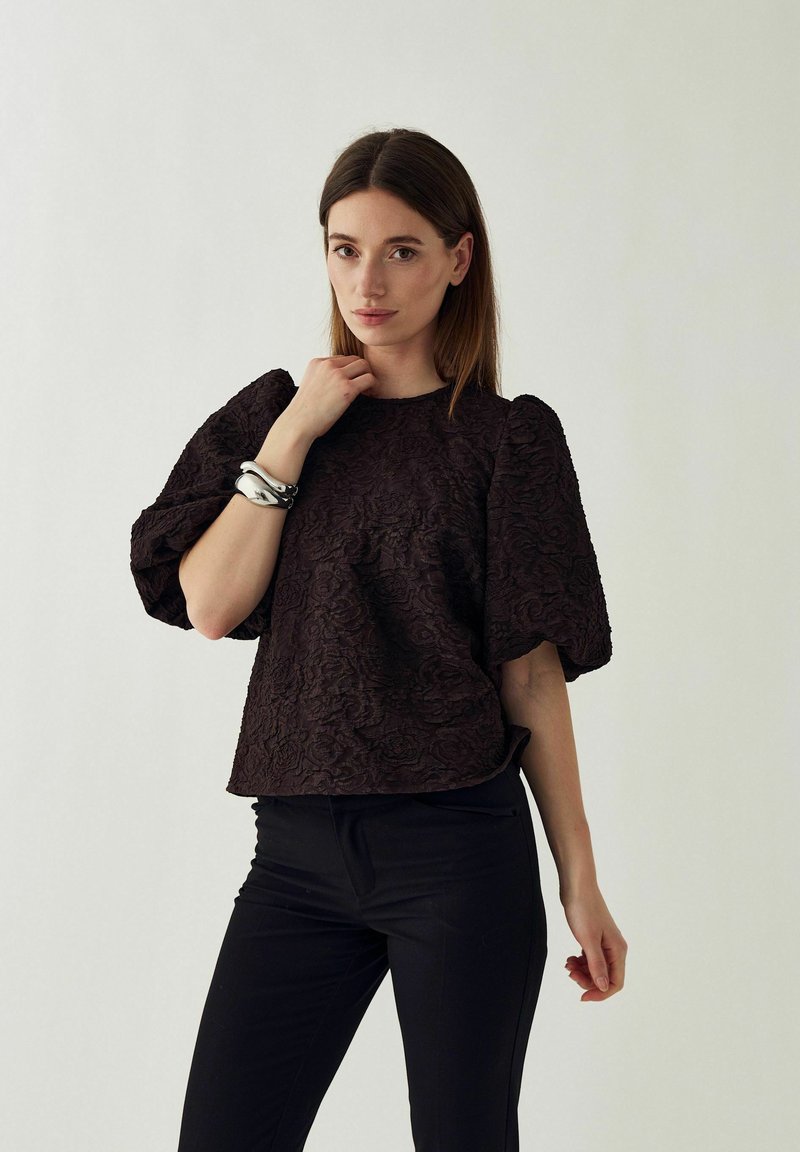 Femme aux cheveux bruns lisses portant un haut sombre texturé à manches bouffantes, un pantalon noir et un bracelet épais en argent, debout à l'intérieur.