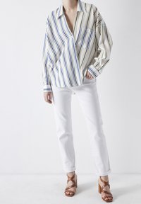 Femme portant une blouse ample à rayures bleues et jaunes, un pantalon blanc retroussé et des sandales à talons beiges, se tenant sur un sol blanc.