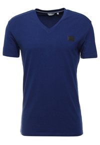 Mörkblå bomulls T-shirt med V-urringning, korta ärmar och en svart rektangulär lapp på bröstet. Slät textur, avslappnad design.