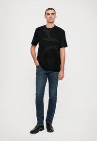 T-shirt preta de manga curta com padrão texturizado, combinada com jeans azul-escuro e sapatos pretos de vestir. O modelo está posicionado contra um fundo simples.