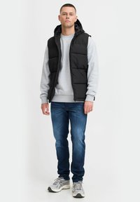 Schwarze gepolsterte Weste mit Kapuze über einem grauen Sweatshirt, kombiniert mit blauen Jeans und grauen Sneakers. Die Weste hat ein горизонтales Steppdesign.