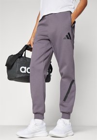 Lilla treningsbukser med svarte detaljer, elastiske mansjetter og sidelommer. Kombinert med hvite joggesko og en svart veske med hvitt logo.