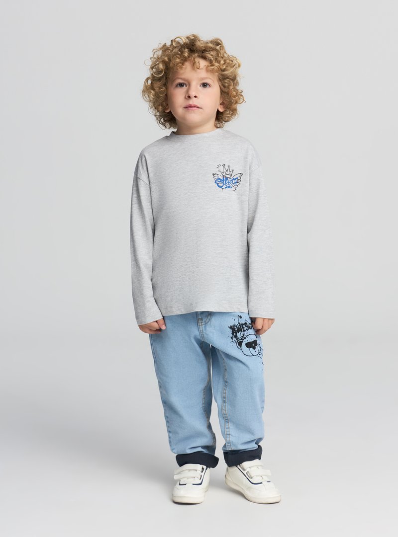 Szara koszula z długim rękawem z niebieskim motylem; jasnoniebieskie spodnie z czarnym nadrukiem; białe trampki na rzepy.