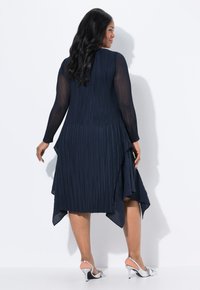Mujer con cabello negro largo, vistiendo un vestido plisado azul marino con mangas transparentes y zapatos de tacón plateados, de pie frente a una pared blanca.