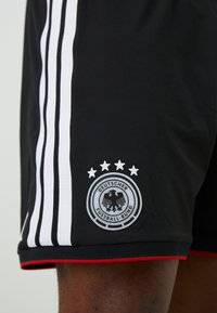 Μαύρα σορτς ποδοσφαίρου με λευκές ρίγες, που διαθέτουν στρογγυλό κεντημένο λογότυπο και τέσσερα αστέρια από πάνω, κόκκινες λεπτομέρειες στο στρίφωμα και υφή υφάσματος.