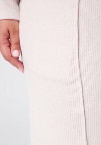 Lichtbeige ribbelstof met een gladde textuur en een zijsplitdetail, met een close-up van het kledingstuk en een hand met gelakte nagels.