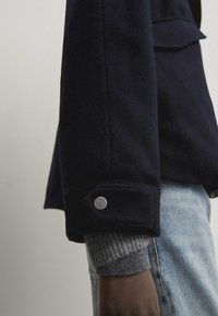 Veste en laine bleu marine à coupe décontractée, avec des boutons argentés aux poignets et un pull gris texturé en dessous, portée avec un jean clair.