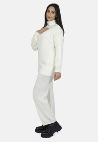 Hvid strikket sweater med høj hals og sideløse kabelmønstre, parret med løstsiddende hvide strikkede bukser og sorte chunky støvler.