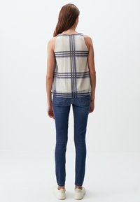 Transparente, ärmellose Bluse mit plissierter Textur und marineblauem Karomuster, kombiniert mit enganliegenden dunklen Jeans und weißen Sneakers.