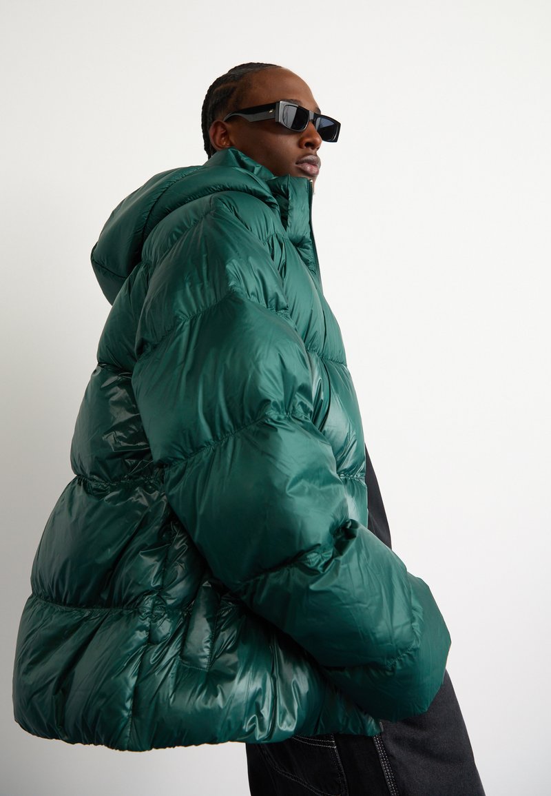 Zielona kurtka puchowa o błyszczącej fakturze, w oversize'owym stylu, z dużym kapturem. Modelka nosi czarne okulary przeciwsłoneczne, zwrócona nieco bokiem.