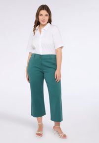 Camicia bianca con colletto a breve, decorata con piccole gemme sul lato sinistro. Pantaloni teal a gamba larga con due bottoni anteriori e texture liscia.