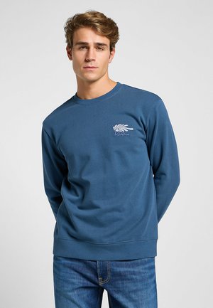 Blå bomull långärmad sweatshirt med ribbad rundhals och fåll. Har en liten vit grafik på vänster bröst. Bärs med blå jeans.