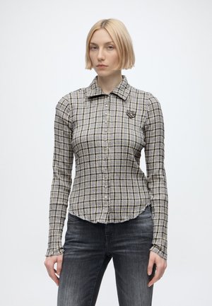Giovane donna con capelli biondi corti che indossa una camicia a quadri con maniche lunghe e jeans grigio scuro, in piedi davanti a uno sfondo semplice.