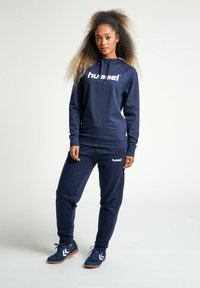 Felpa atletica blu navy e pantaloni realizzati in tessuto liscio, con un logo bianco ben visibile, dotati di coulisse e polsini a coste.