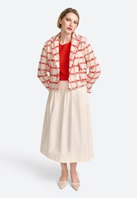 Veste à carreaux rouge et blanc texturée sur un haut rouge, accompagnée d'une longue jupe couleur crème et de talons nude, avec une coupe ample.