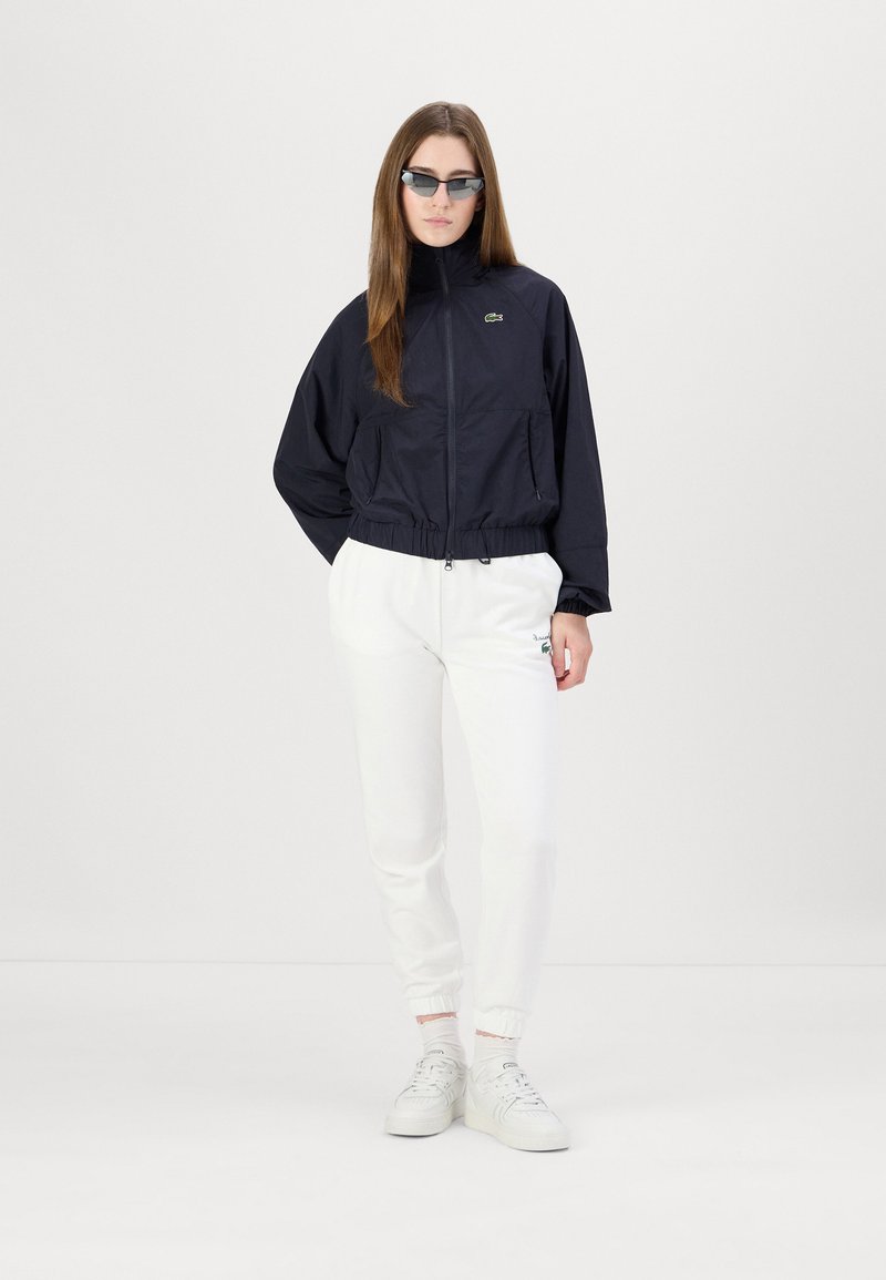 Veste légère noire zippée avec des poignets élastiques, associée à un pantalon de jogging blanc arborant un petit logo vert. Les accessoires comprennent des lunettes de soleil rectangulaires de petite taille.