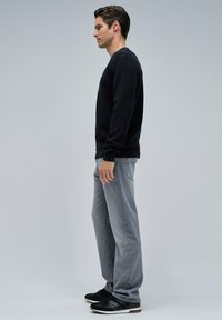 Suéter negro, jeans grises, zapatillas negras con acentos texturizados. Perfil lateral que muestra el ajuste y el estilo, contra un fondo liso.