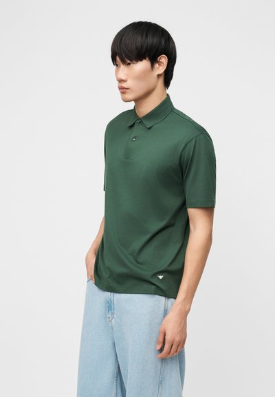 Jeune homme aux cheveux noirs portant un polo à manches courtes vert foncé et un jean bleu clair, debout les mains dans les poches.