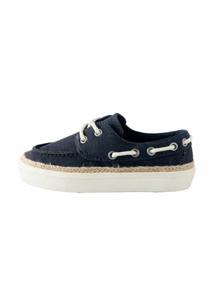 Scarpa da barca in tela blu navy con lacci bianchi, dettaglio in corda sopra la suola bianca in gomma e occhielli metallici sul lato e sul davanti.