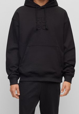 Schwarzer Hoodie mit Fronttasche und Kordelzug, dezentes, geprägt "HUGO"-Logo auf der Brust, getragen mit passenden schwarzen Hosen.