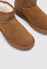 Botines de ante marrón con un forro suave beige, suelas planas de goma, y diseño de punta redondeada. Presentan detalles de costura a lo largo de las costuras.