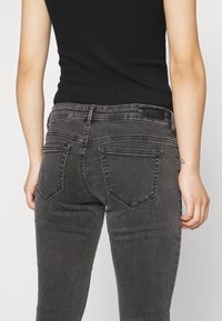 Persona che indossa jeans neri sbiaditi e un top senza maniche nero, mostrando la schiena con un piccolo tatuaggio a forma di cuore sul polso destro.