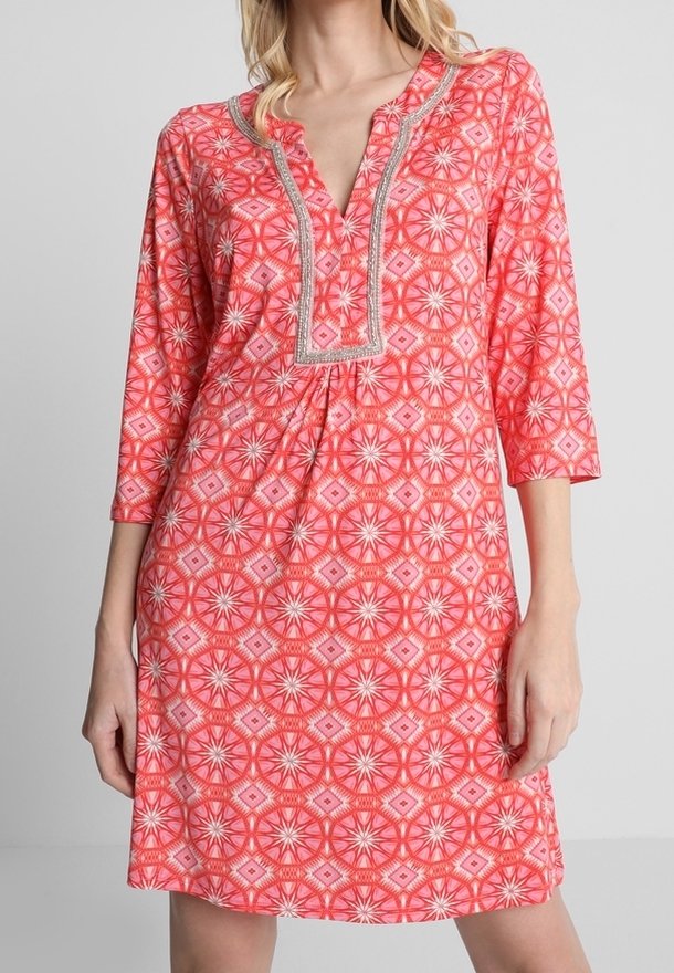 Femme portant une robe corail rose longueur genoux avec des manches trois quarts et des motifs géométriques en forme d'étoiles, dotée d'un décolleté en V orné d'une bordure argentée.