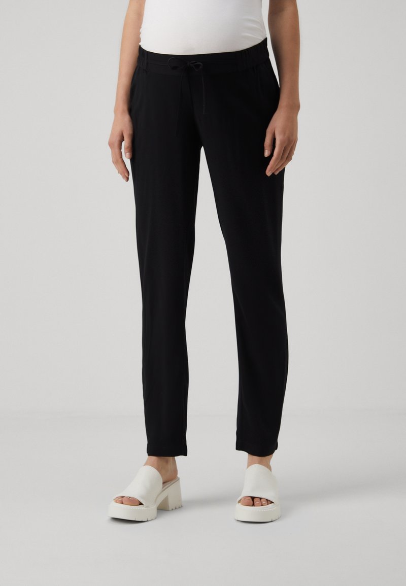 Femme portant un pantalon noir à cordon et des sandales blanches à talons et orteils ouverts, debout devant un fond clair uni.