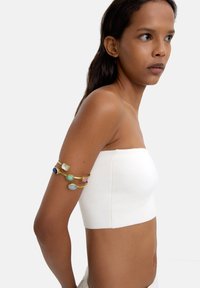 Złotonośny metalowy rękaw z wieloma kolorowymi kamieniami szlachetnymi: niebieskim, zielonym, różowym i przezroczystym. Noszony na nagiej ręce obok białego topu bez ramiączek.