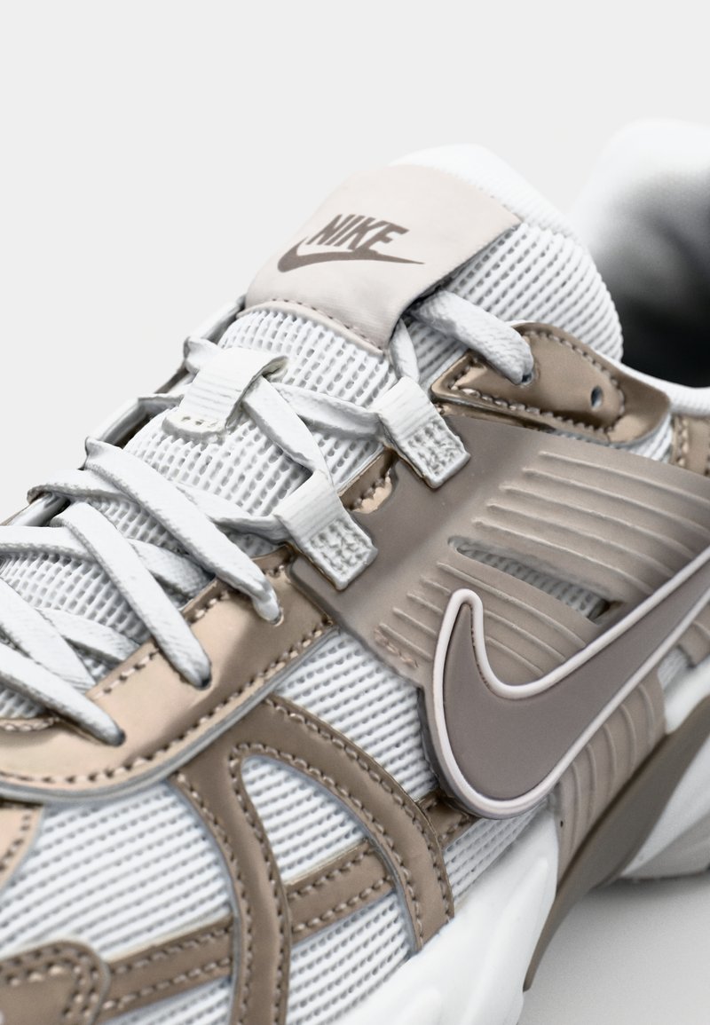 Zapatilla de malla blanca con acentos beige, que presenta un diseño texturizado, un prominente logo de Nike y cierre con cordones. Los detalles incluyen patrones cosidos.