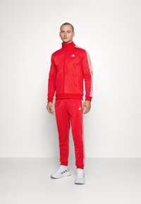 Rdeč trenirka iz gladkega materiala ima bele tri stripe na rokavih in hlačah ter Adidas logo na prsih in boku.