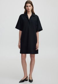 Abito nero a maniche corte con colletto, forma rilassata e tessuto liscio. Un paio di scarpe slingback nere completa l'outfit.