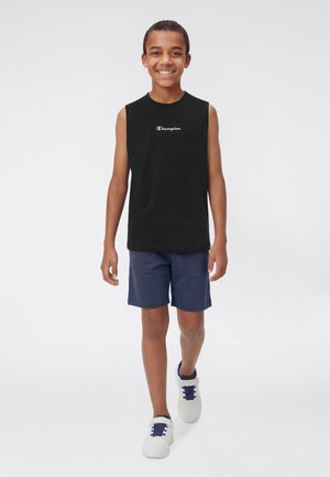 Ragazzo sorridente che cammina in avanti indossando una canotta nera Champion, pantaloncini blu navy e scarpe da ginnastica bianche con lacci blu su uno sfondo bianco.