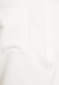 Chemise blanche avec une poche plaquée, texture lisse et patte de boutonnage cachée. Le tissu semble léger avec un léger brillant.